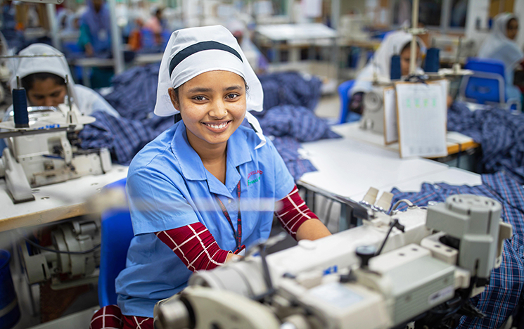 A Bangladeshi woman sits at a sewing machine, smiling at the camera.